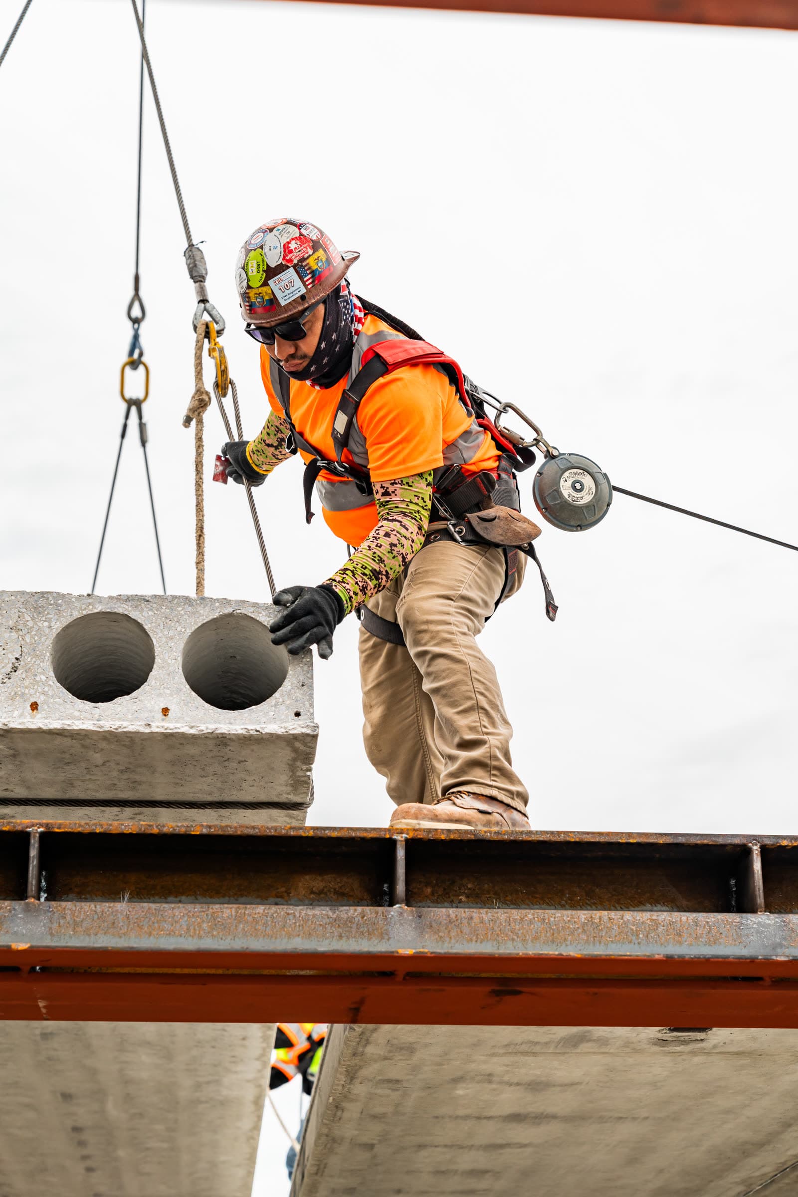 Worker with fall-arrest reel guiding a hollow-core plank onto a steel beam